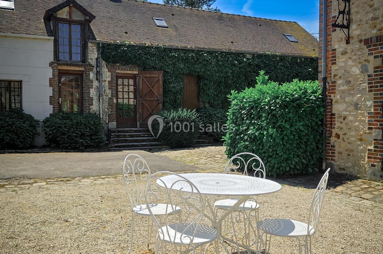 Table et chaises blanches en fer forgé sur une cour en gravier, devant une maison en pierre avec façade végétalisée.