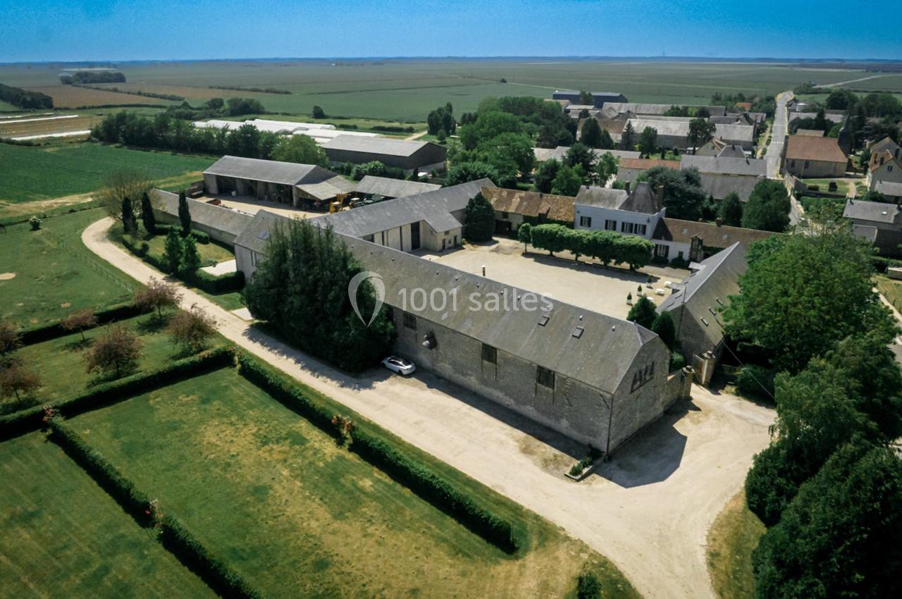 Vue aérienne d'une ferme avec bâtiments en pierre, cour centrale et jardins entourés de champs et d'habitations rurales.