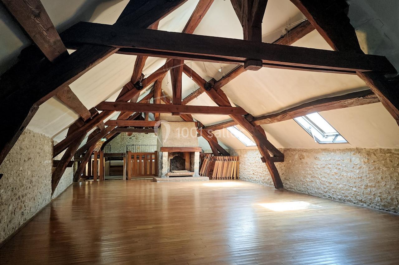 Grande pièce sous combles avec poutres apparentes, parquet en bois, murs en pierre et cheminée centrale.