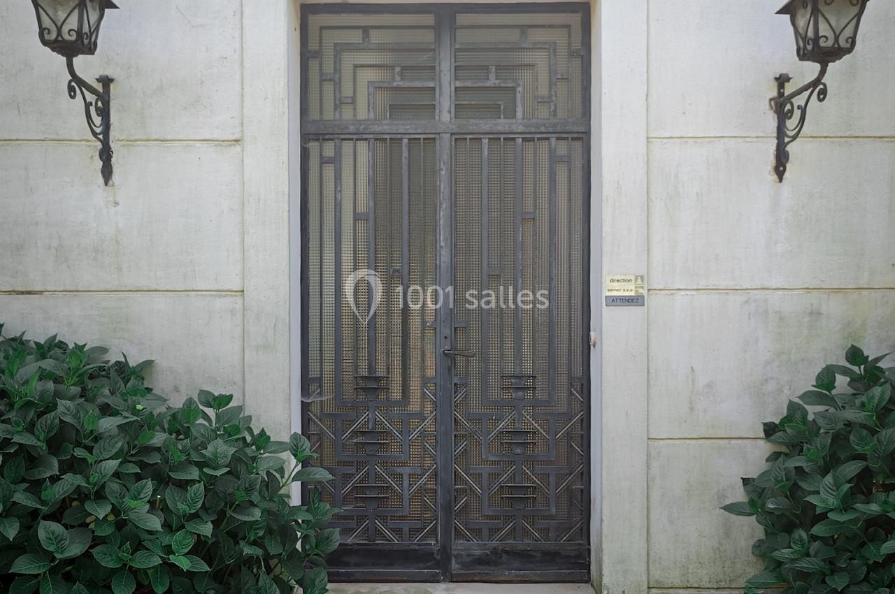 Porte en fer forgé au design géométrique, encadrée par deux lampes murales et des arbustes sur un sol pavé.
