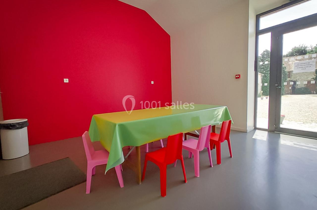 Table recouverte d'une nappe verte entourée de petites chaises colorées dans une pièce aux murs rouges et blancs.