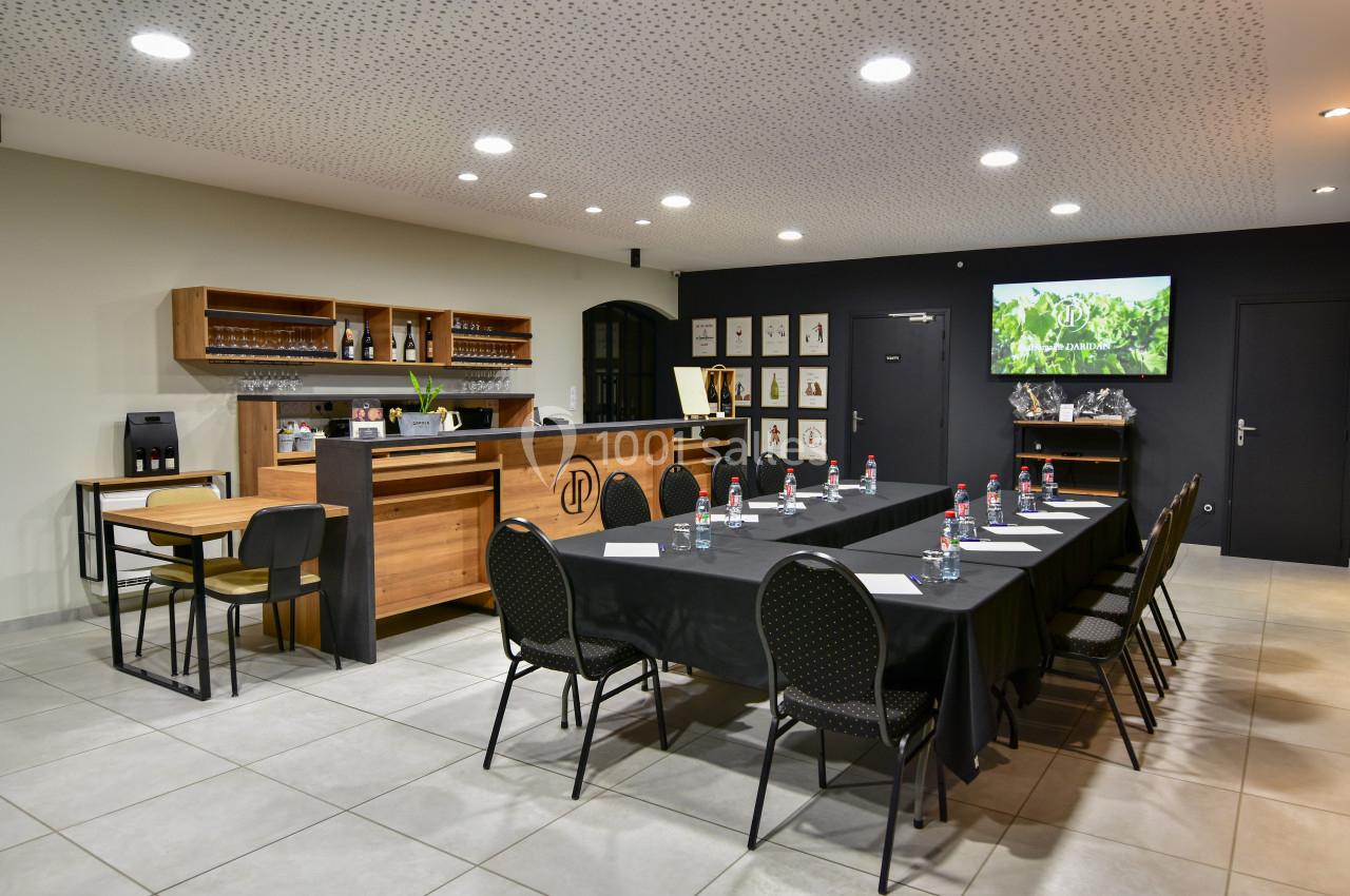 Salle de réunion avec une grande table rectangulaire, chaises noires, écran mural et coin bar en arrière-plan.