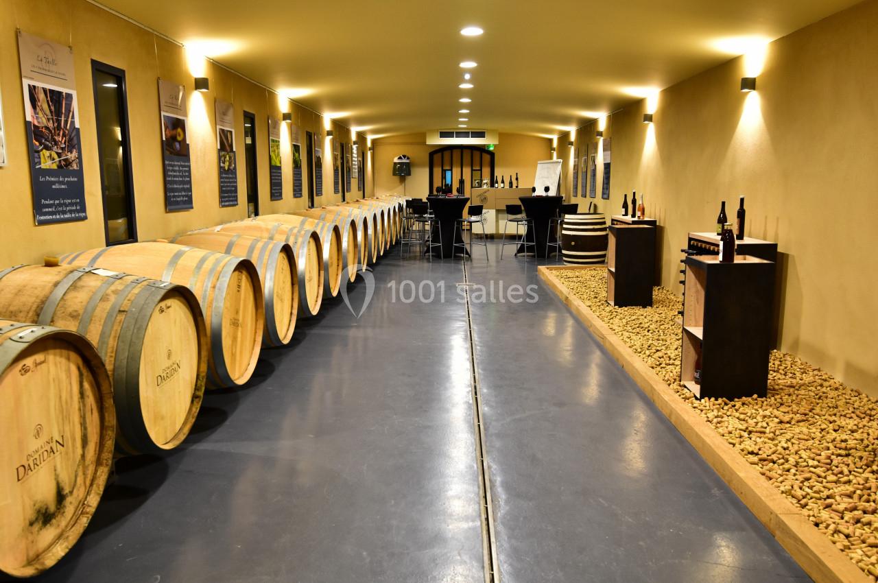 Salle de dégustation avec des tonneaux alignés à gauche, des bouteilles exposées à droite et un espace aménagé au fond.