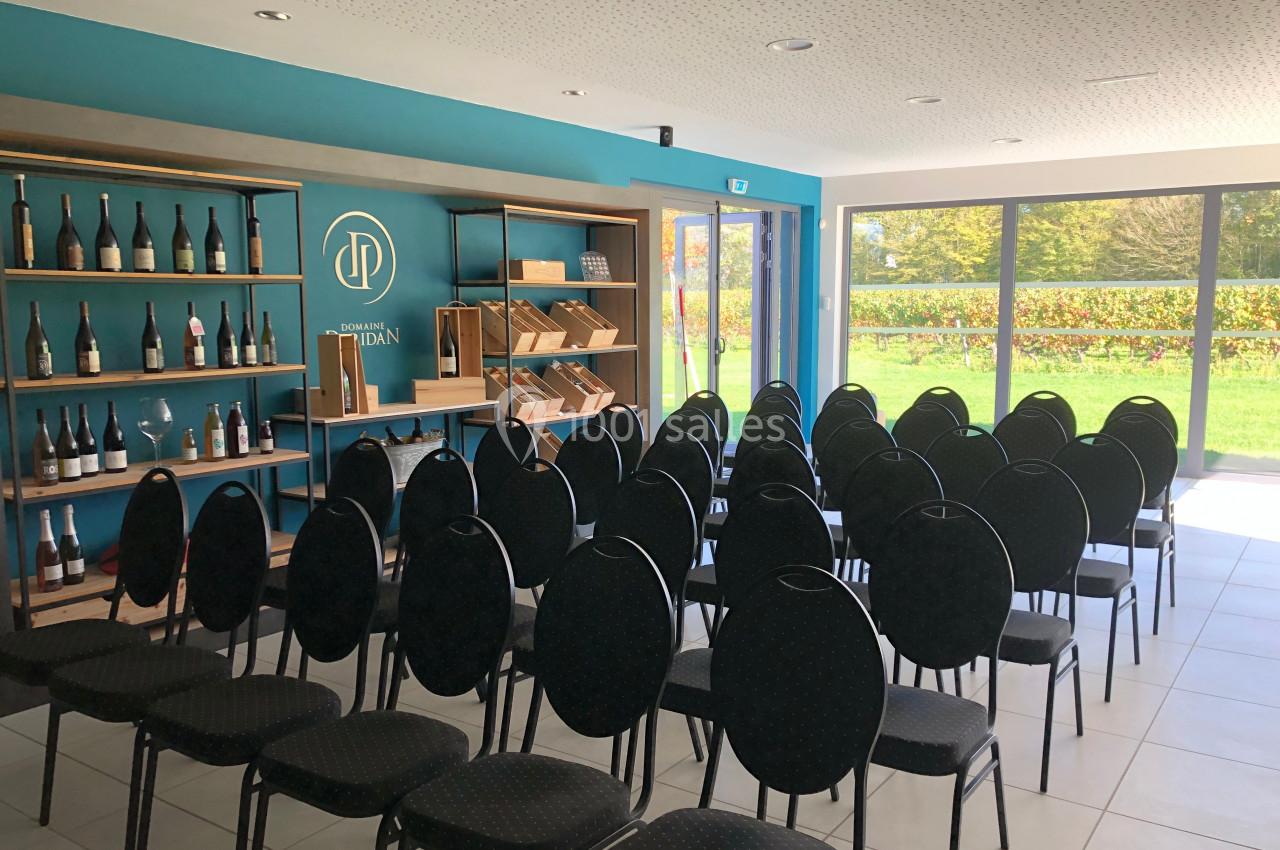 Salle lumineuse avec des chaises alignées face à une étagère contenant des bouteilles et des livres, vue sur un jardin.