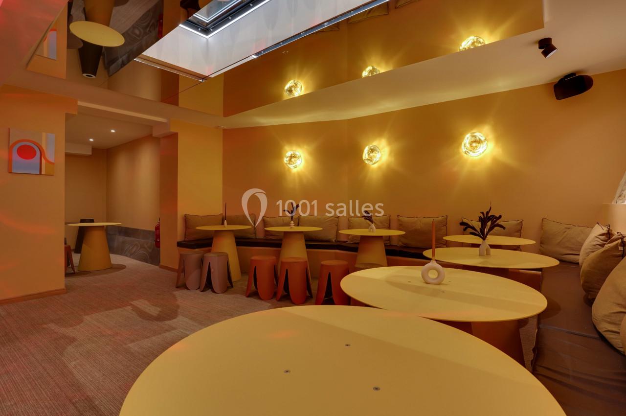 Salle lumineuse avec murs jaunes, tables rondes assorties et banquettes garnies de coussins sous des appliques murales.