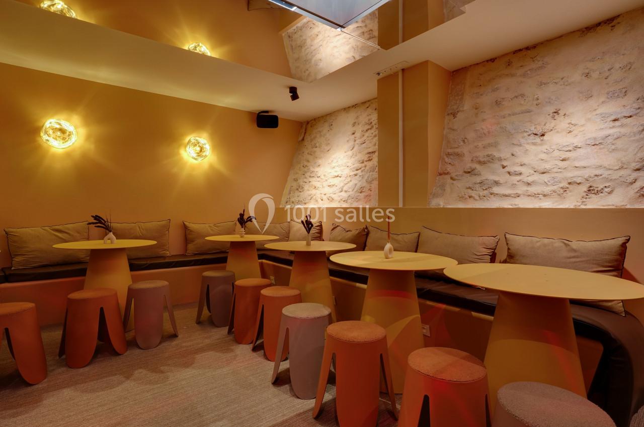 Salle lumineuse avec murs en pierre, tables rondes jaunes, tabourets assortis et éclairage mural chaleureux.