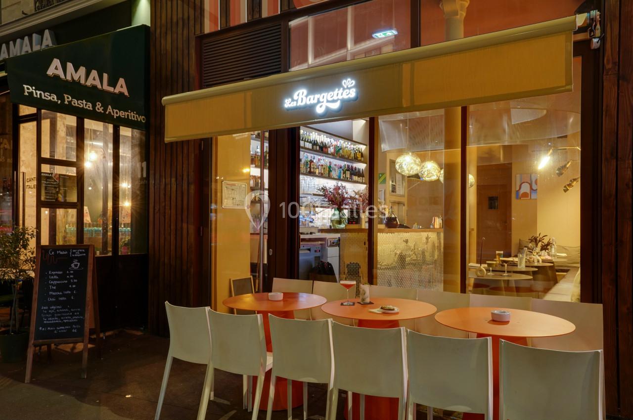 Façade d'un restaurant avec terrasse extérieure, tables et chaises, éclairage chaleureux en soirée.