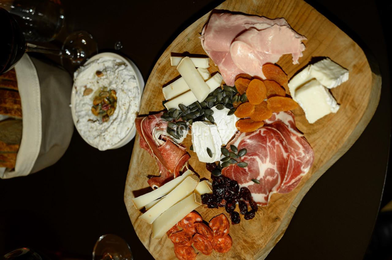 Plateau de charcuterie et fromages variés avec fruits secs, graines et pain en arrière-plan sur une table sombre.