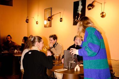 Une personne de dos prépare un cocktail avec un shaker dans un bar, portant un t-shirt blanc à motif orange.
