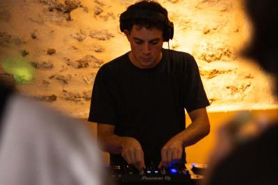 Une personne de dos prépare un cocktail avec un shaker dans un bar, portant un t-shirt blanc à motif orange.