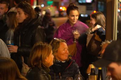 Une personne de dos prépare un cocktail avec un shaker dans un bar, portant un t-shirt blanc à motif orange.