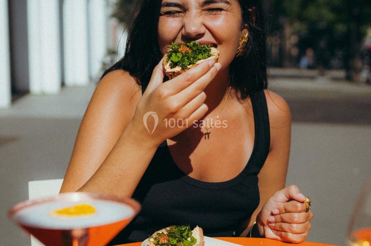 Femme souriante mangeant une tartine garnie de légumes verts à une table en extérieur par une journée ensoleillée.