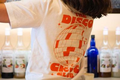 Une personne de dos prépare un cocktail avec un shaker dans un bar, portant un t-shirt blanc à motif orange.