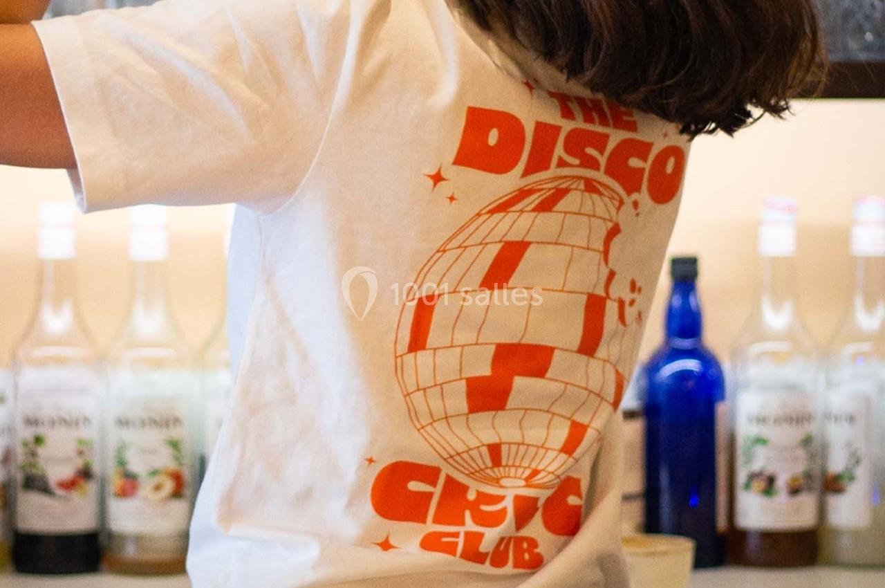 Une personne de dos prépare un cocktail avec un shaker dans un bar, portant un t-shirt blanc à motif orange.