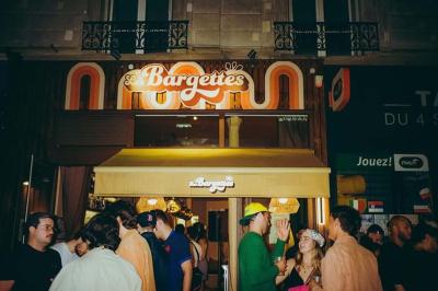 Une personne de dos prépare un cocktail avec un shaker dans un bar, portant un t-shirt blanc à motif orange.