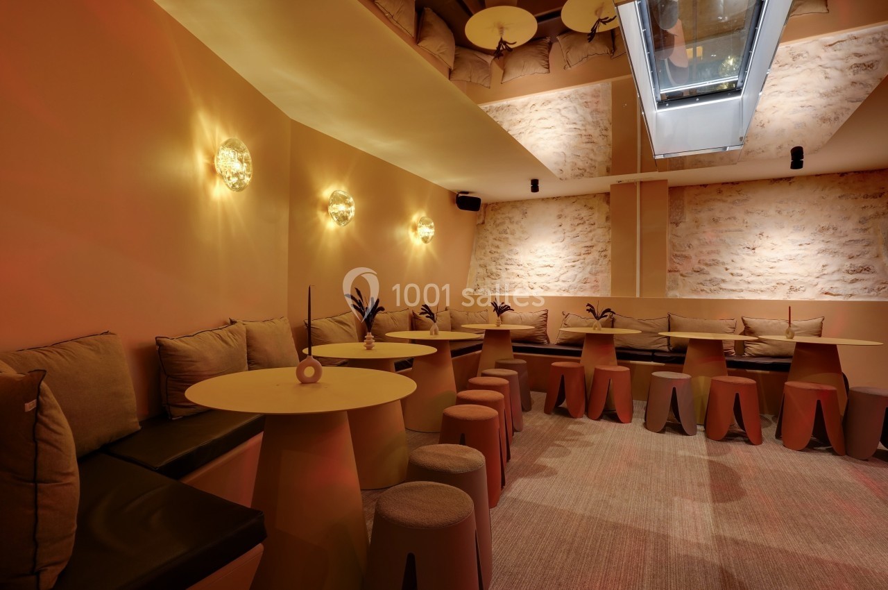 Salle lumineuse avec murs en pierre, tables rondes, tabourets et banquettes beige sous un puits de lumière.