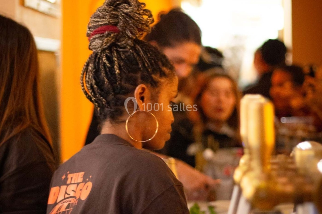 Une femme de dos avec des tresses et des boucles d'oreilles se tient près d'un comptoir dans un lieu animé.