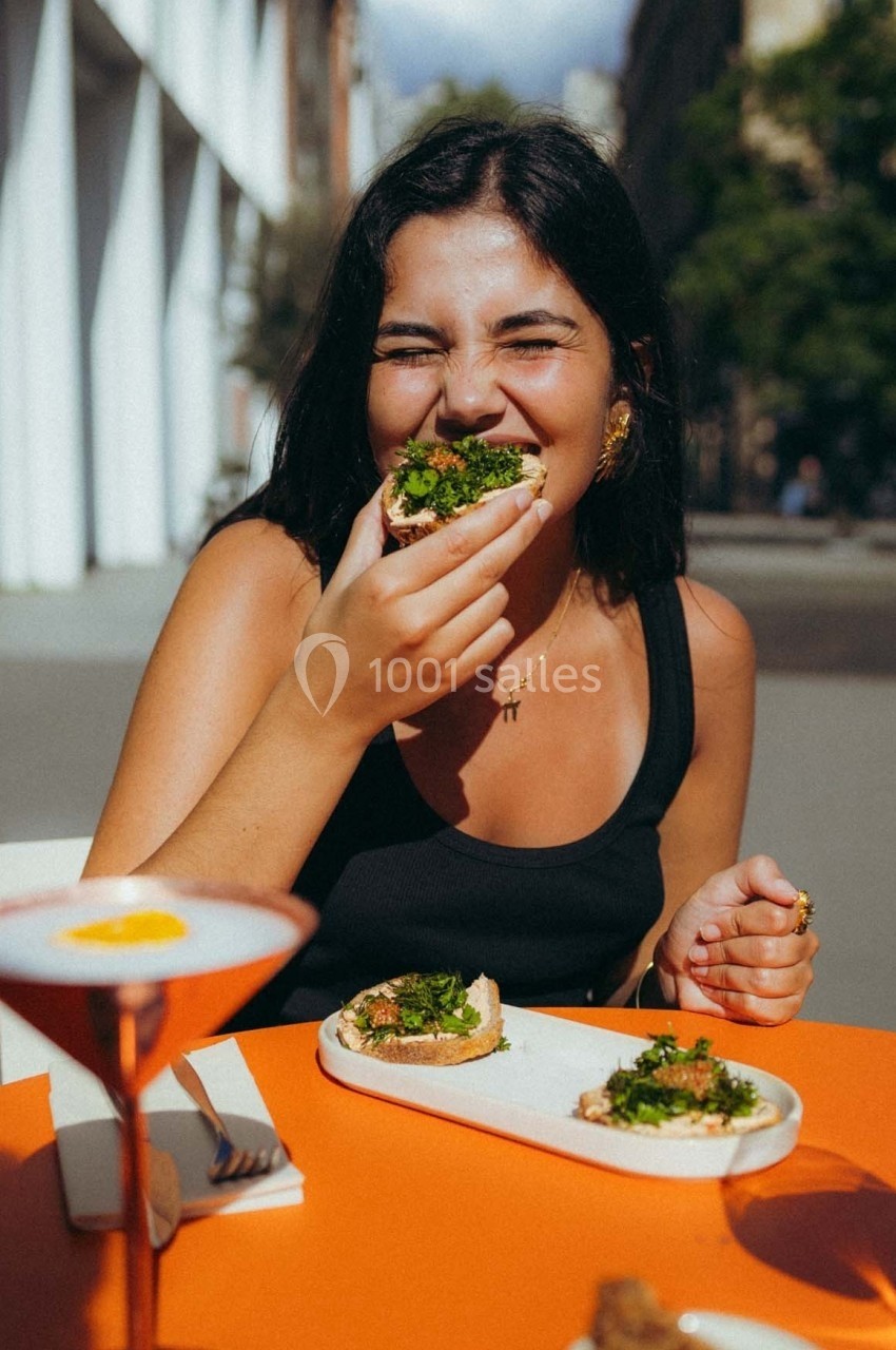 Une femme souriante mange une tartine garnie de légumes verts à une table en extérieur par une journée ensoleillée.