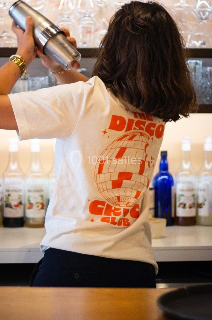 Une personne de dos en train de secouer un shaker derrière un bar, portant un t-shirt avec un motif disco.