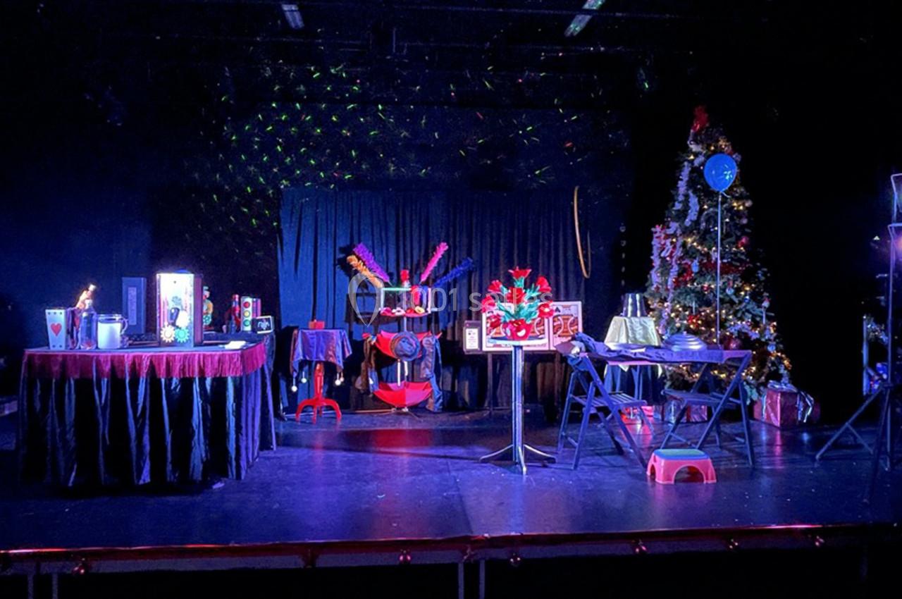 Scène de spectacle avec décorations festives, rideaux noirs, tables ornées et un sapin de Noël illuminé.