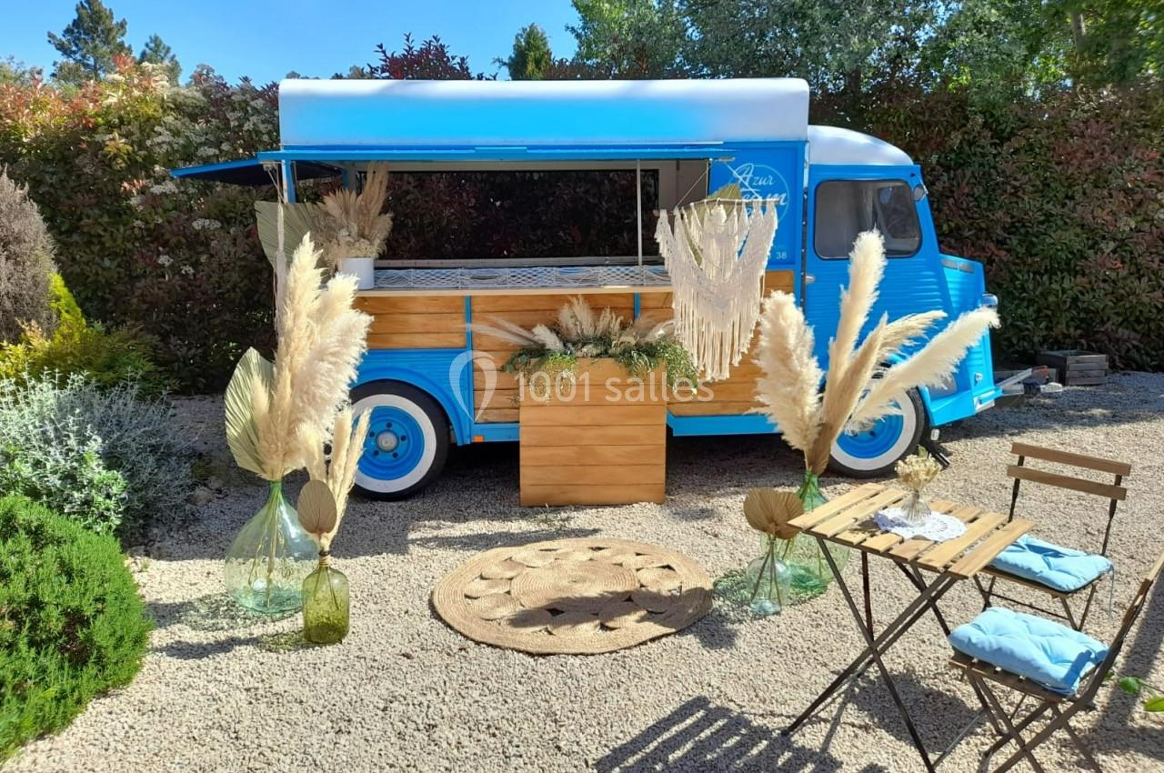 Camion bleu aménagé en food truck, entouré de décorations en pampas, avec table et chaises en bois sur un sol gravillonné.