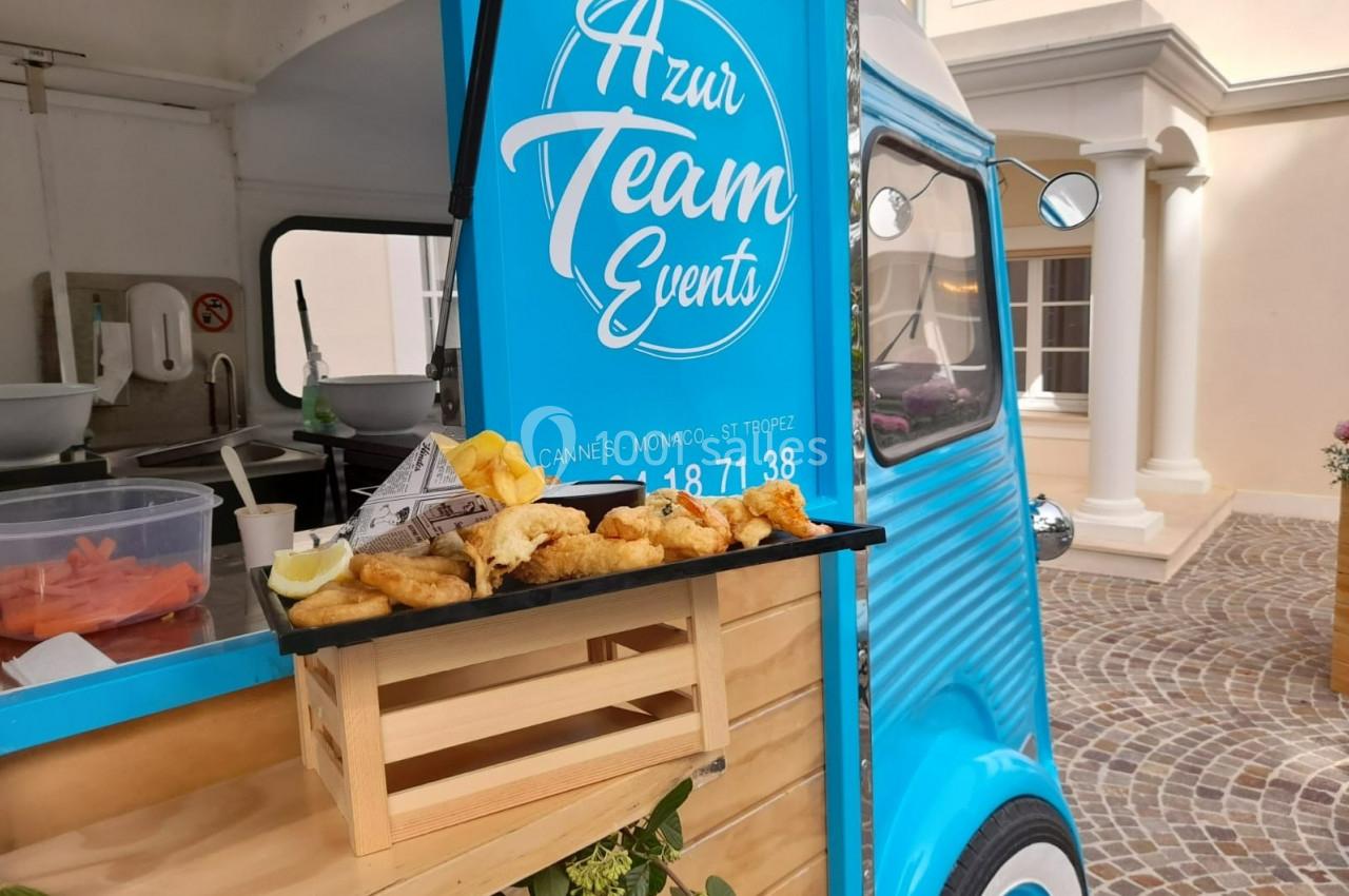 Plateau de beignets dorés posé sur une petite caisse en bois devant un food truck bleu stationné.