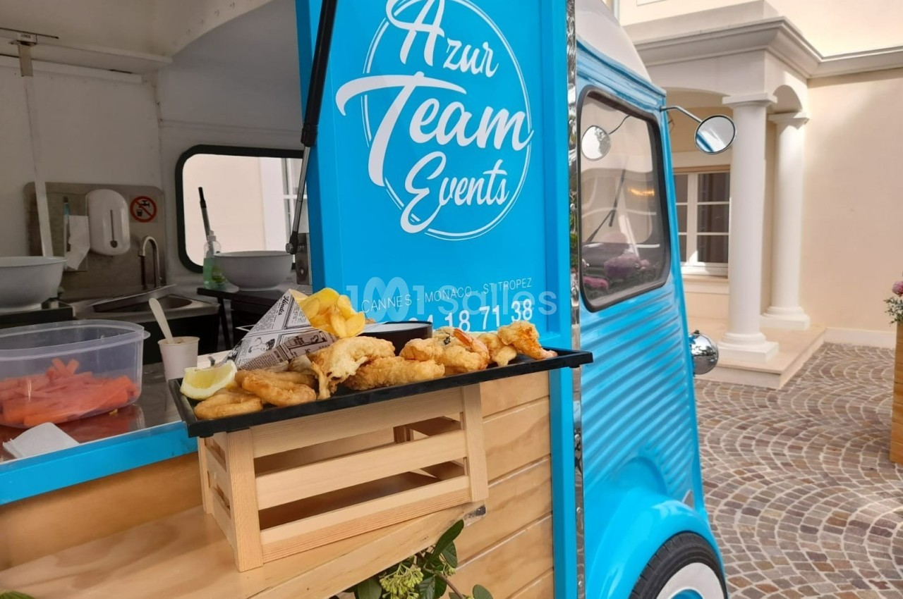 Plateau de beignets dorés posé sur une petite caisse en bois devant un food truck bleu stationné.