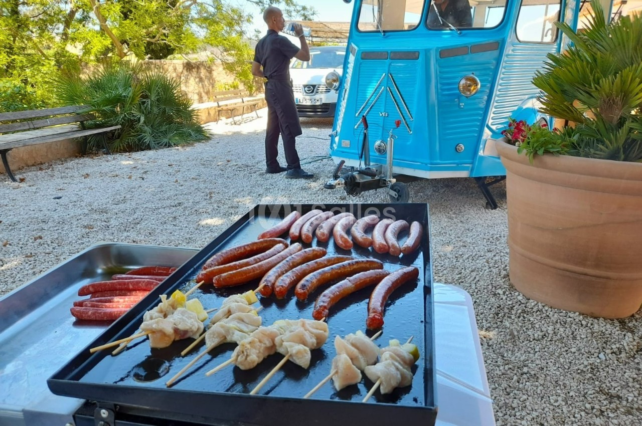 Plancha avec brochettes de poulet et saucisses grillées, devant un food truck bleu dans une cour ensoleillée.