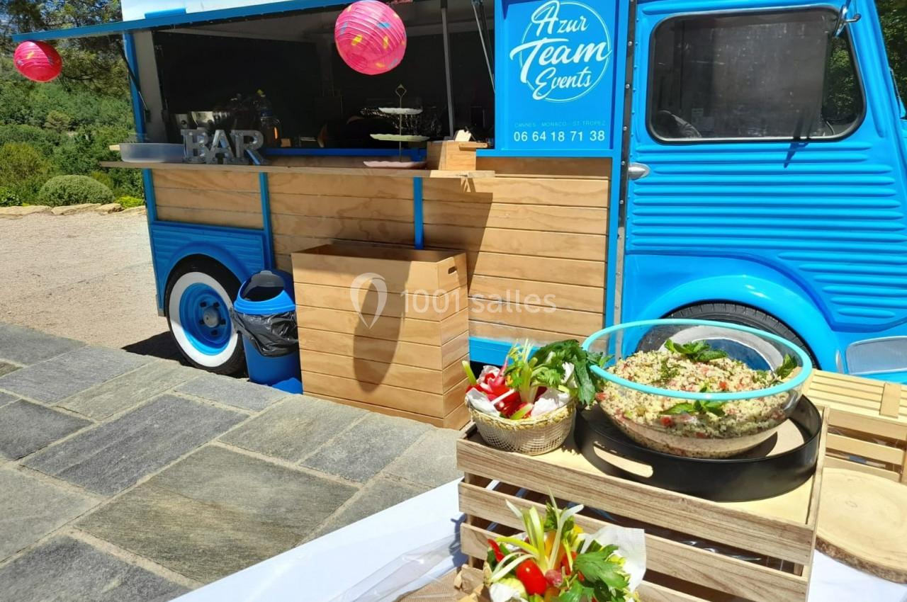Un food truck bleu stationné en extérieur avec des plats frais disposés sur des caisses en bois au premier plan.
