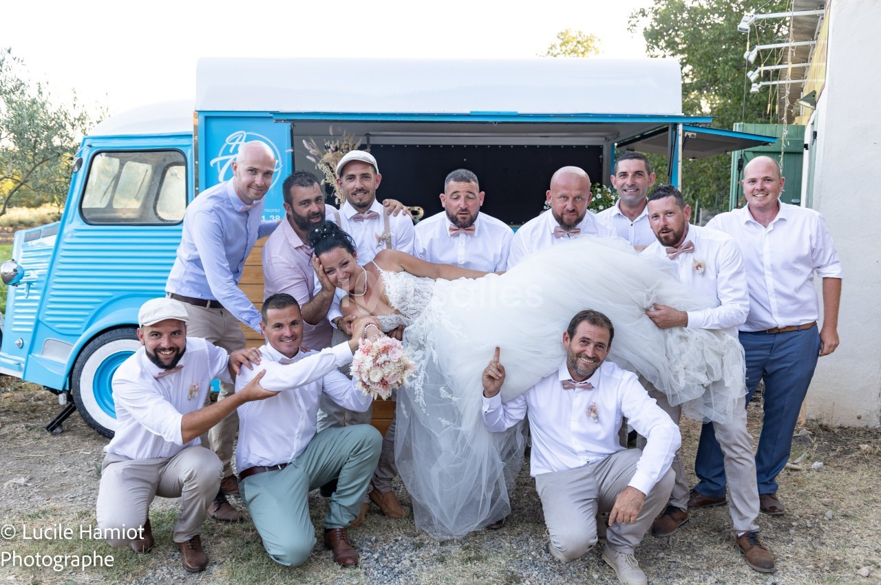 Un groupe d'hommes en chemises blanches porte une mariée souriante devant un camion bleu vintage.
