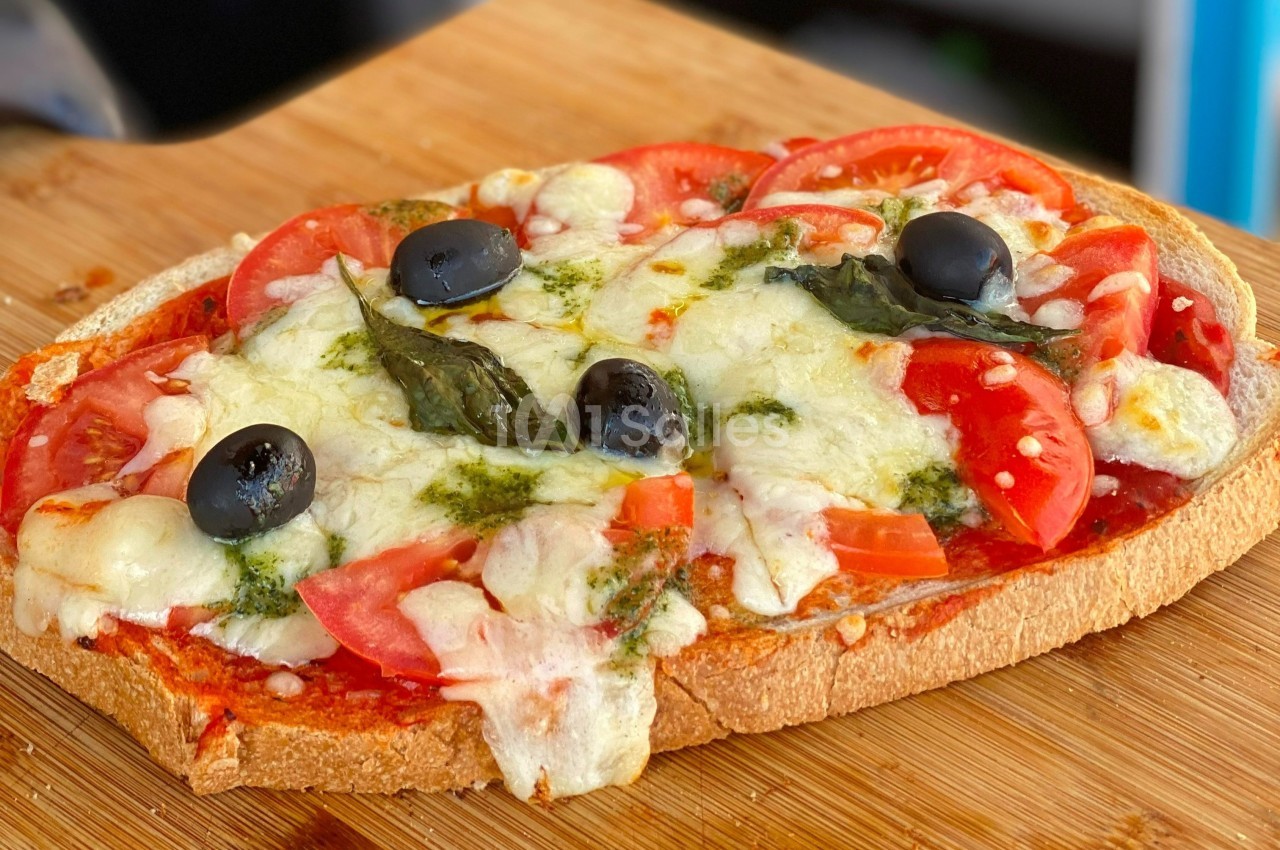 Tranche de pain garnie de tomates, mozzarella fondue, olives noires, basilic et pesto, posée sur une planche en bois.