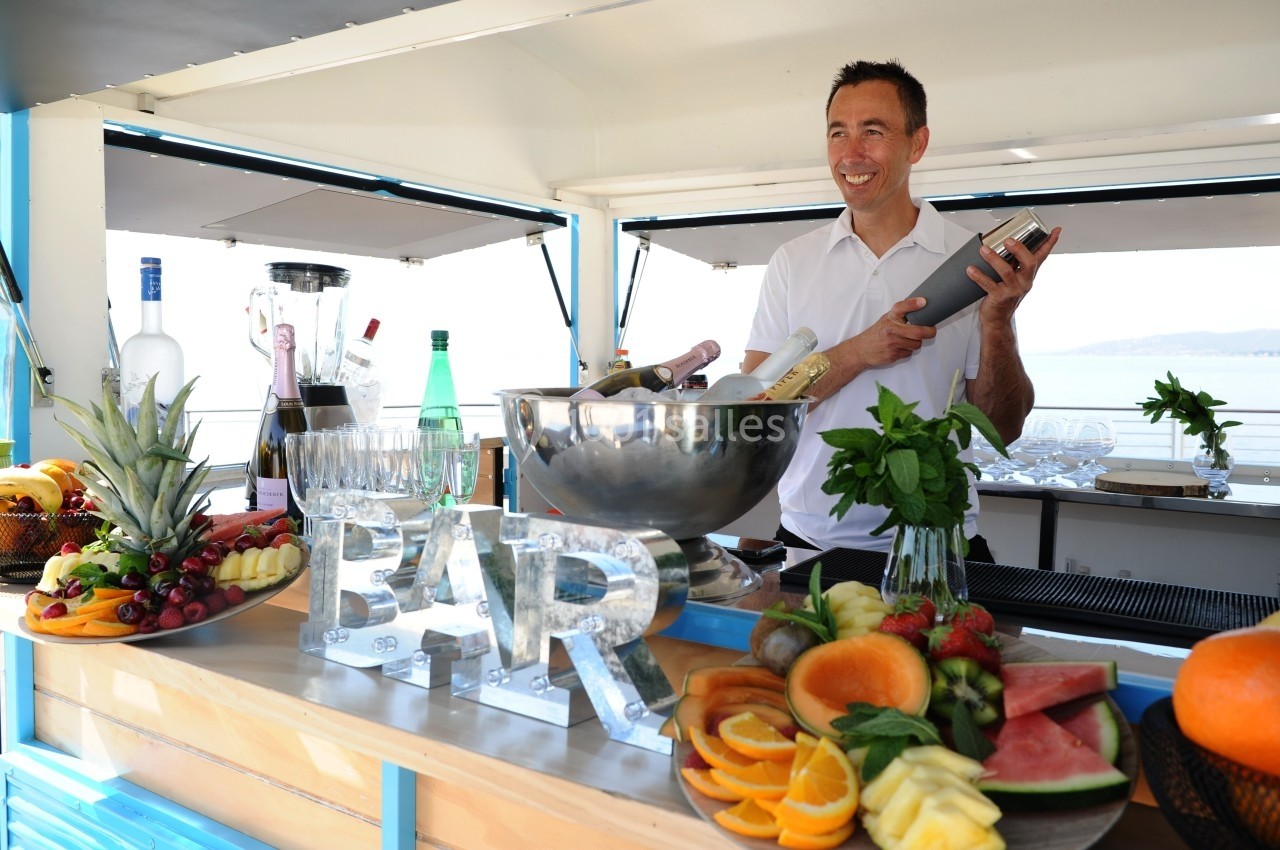 Un barman souriant prépare un cocktail derrière un bar garni de fruits frais et de bouteilles.