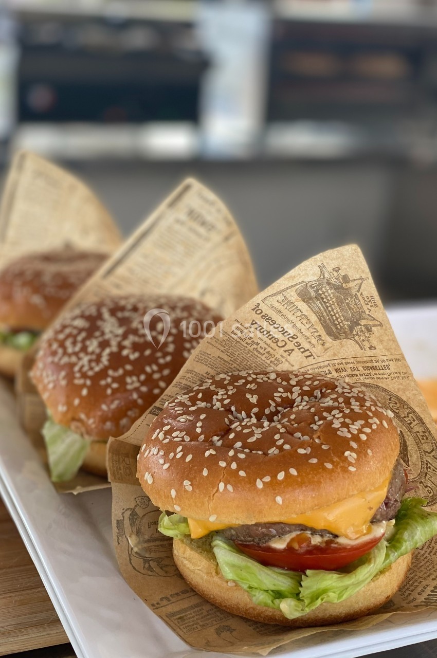 Trois hamburgers garnis de salade, tomate et fromage, enveloppés dans du papier imprimé, présentés sur un plateau blanc.