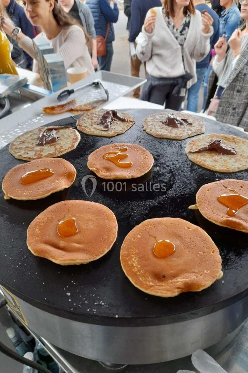 Crêpes et pancakes en cours de cuisson sur une plaque chauffante, garnis de confiture et de chocolat.