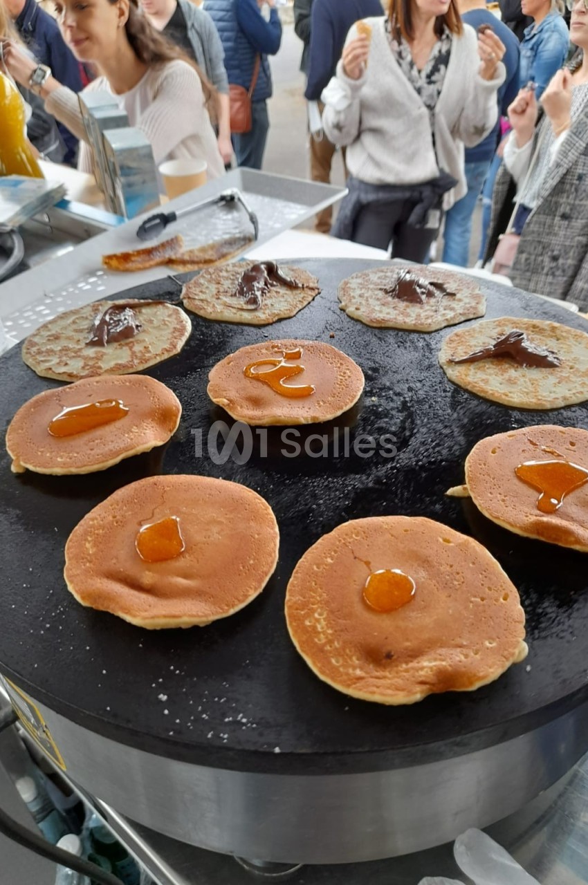 Crêpes et pancakes en cours de cuisson sur une plaque chauffante, garnis de confiture et de chocolat.