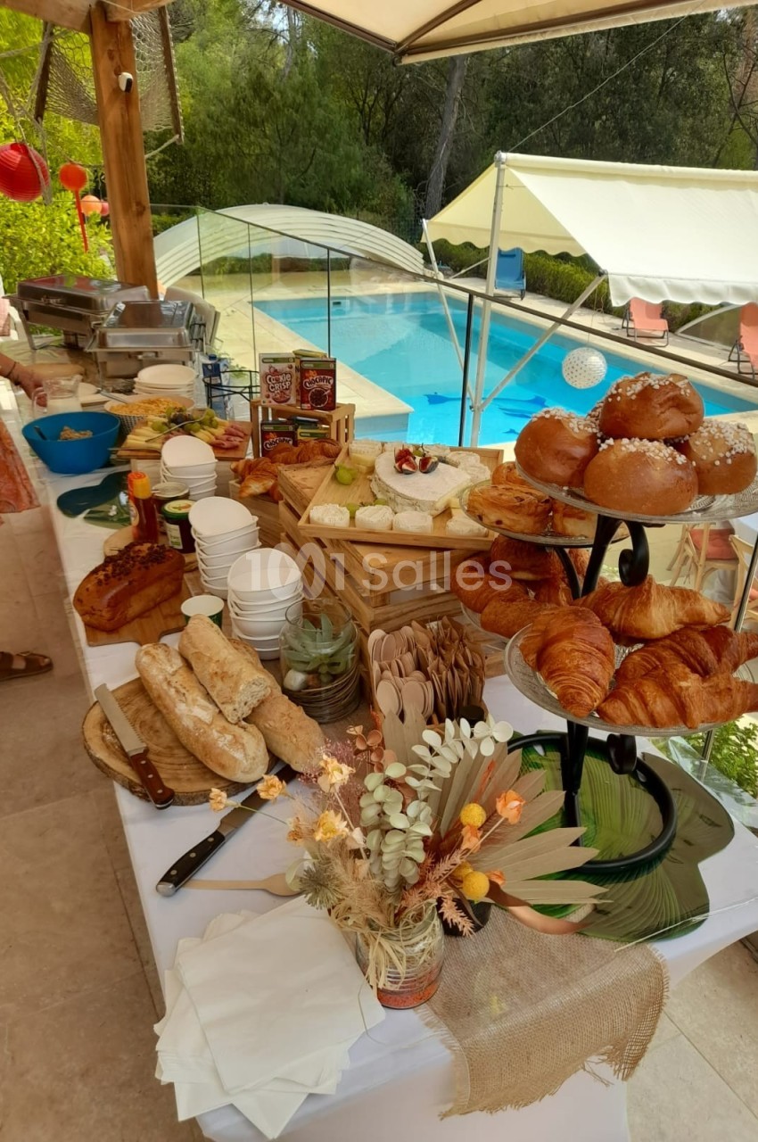 Buffet en extérieur avec viennoiseries, pains, fromages et fruits, près d'une piscine entourée de verdure.