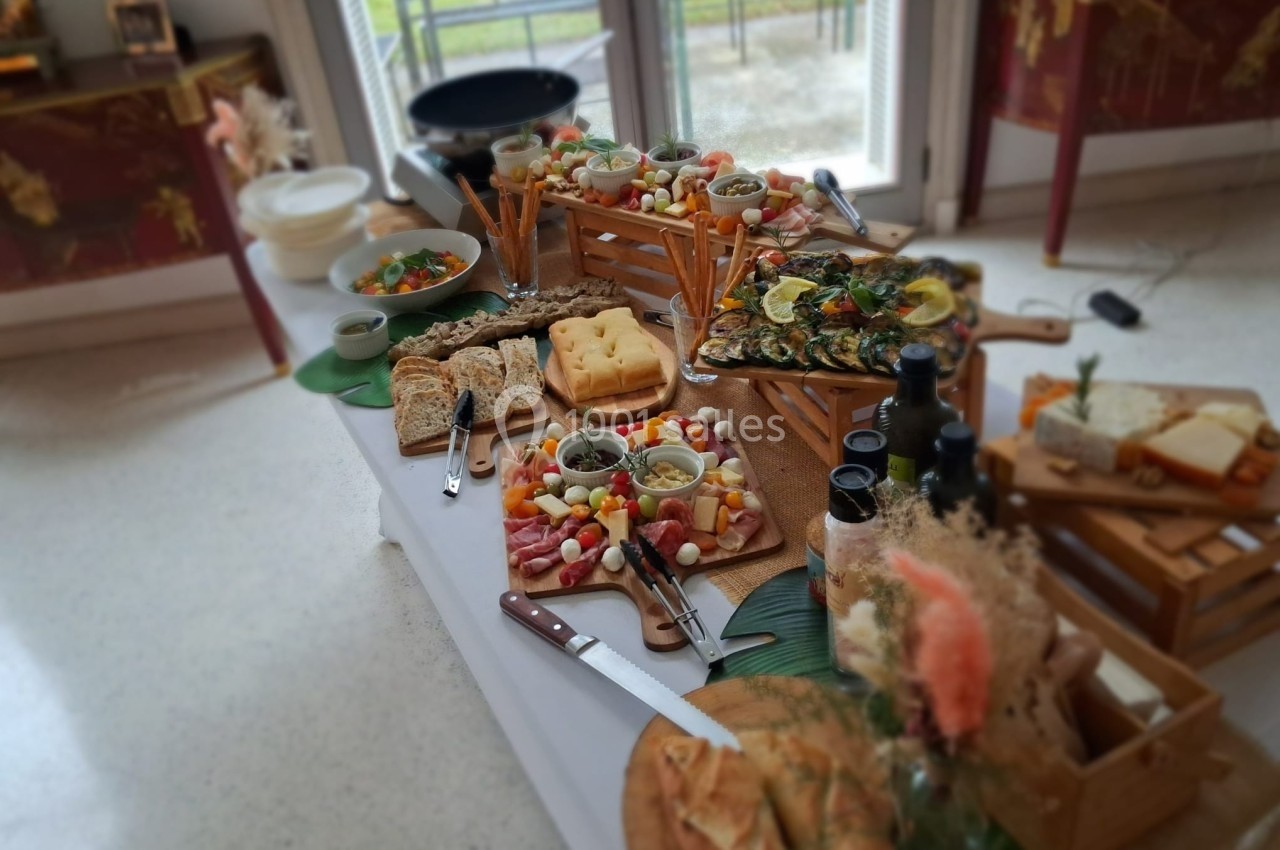Table garnie de plats variés, incluant charcuterie, légumes, pains et fromages, dans un cadre lumineux près d'une fenêtre.