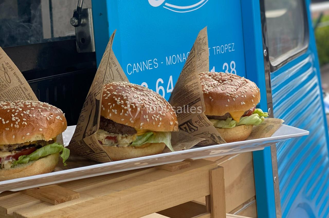 Trois hamburgers garnis de salade, fromage et steak, présentés sur un plateau devant un food truck bleu.