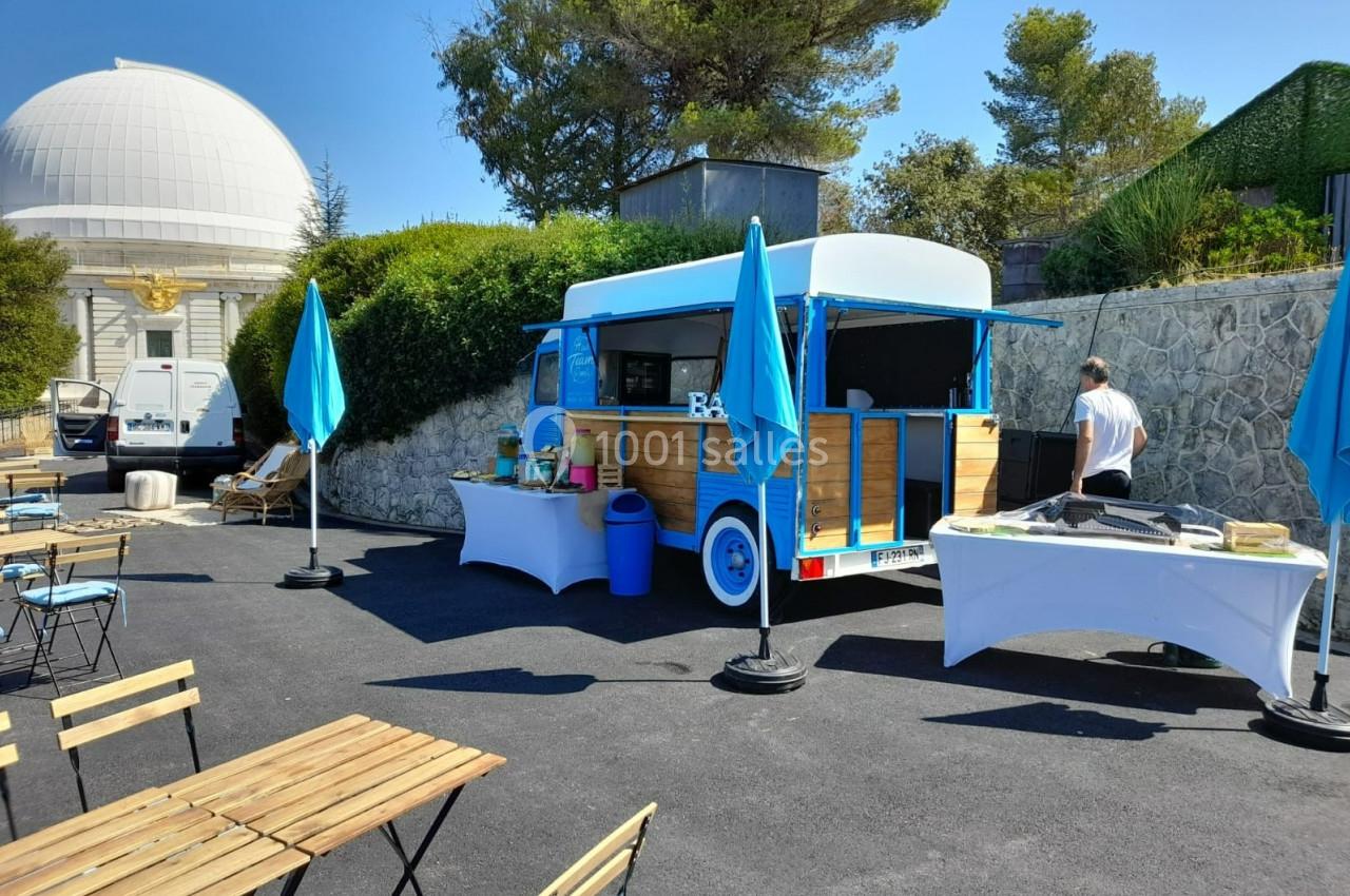 Un food truck en bois et bleu avec des tables et parasols installés sur un espace extérieur pavé.
