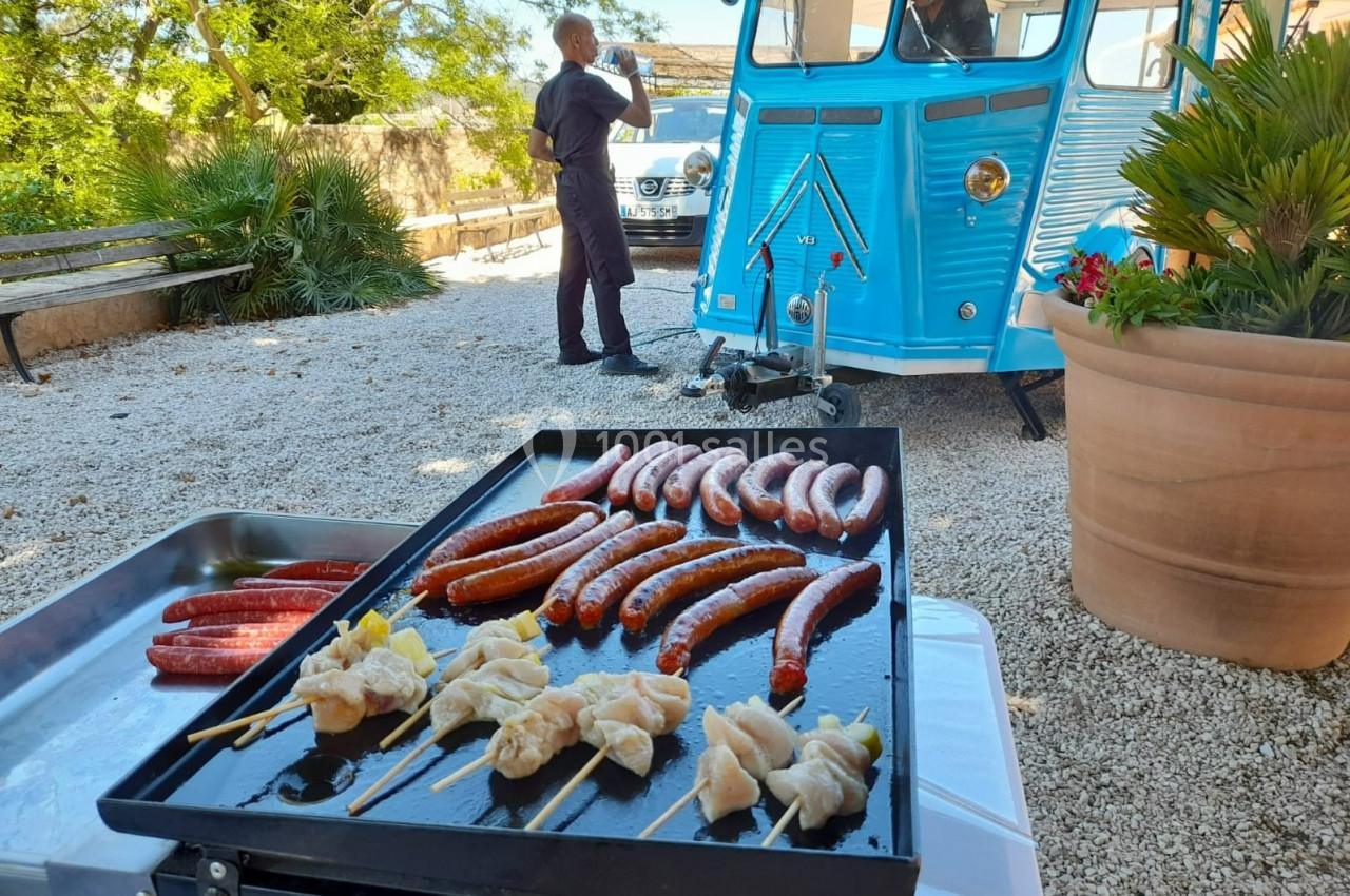 Plancha avec brochettes de poulet et saucisses grillées, devant un food truck bleu dans une cour ensoleillée.
