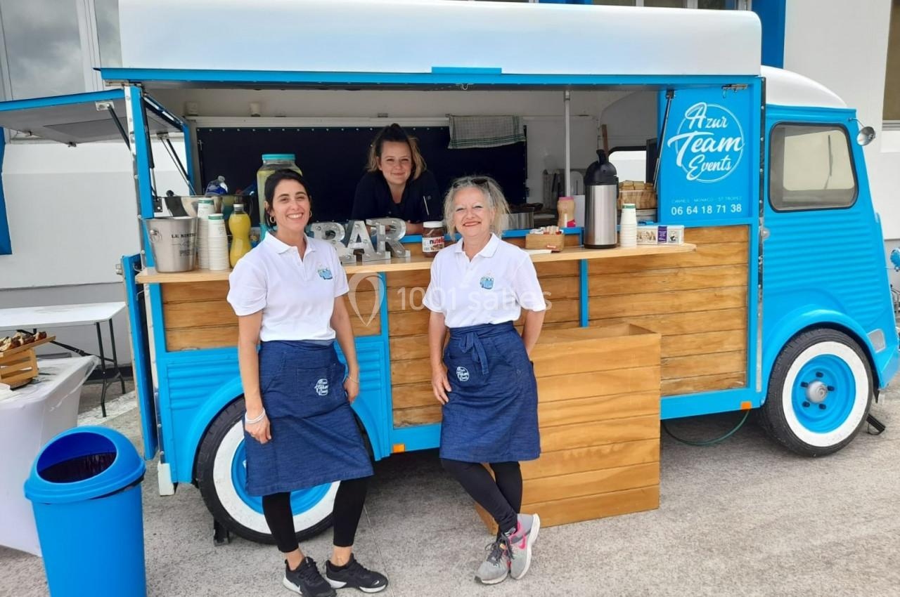 Un food truck bleu avec trois personnes souriantes en tablier devant un comptoir en bois.