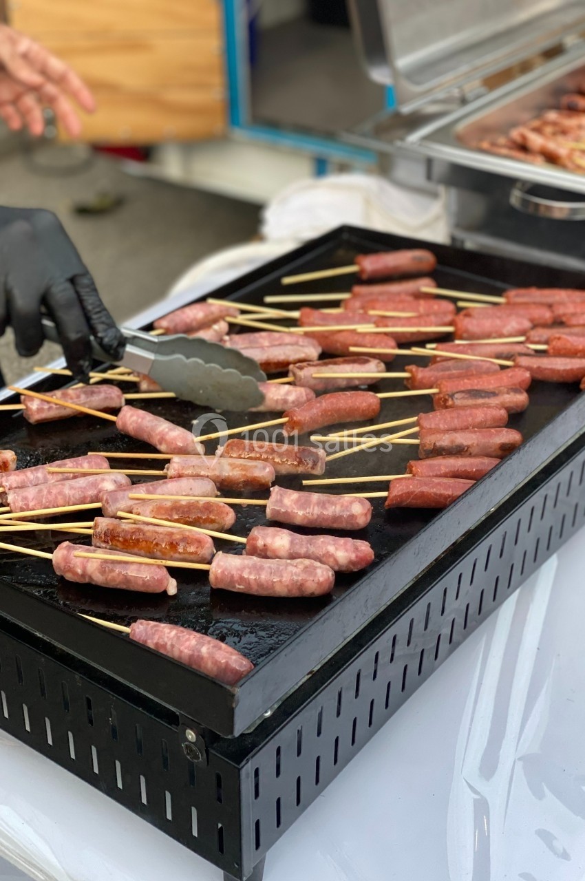 Saucisses sur brochettes grillées sur une plaque chauffante, manipulées avec des pinces par une personne portant des gants…