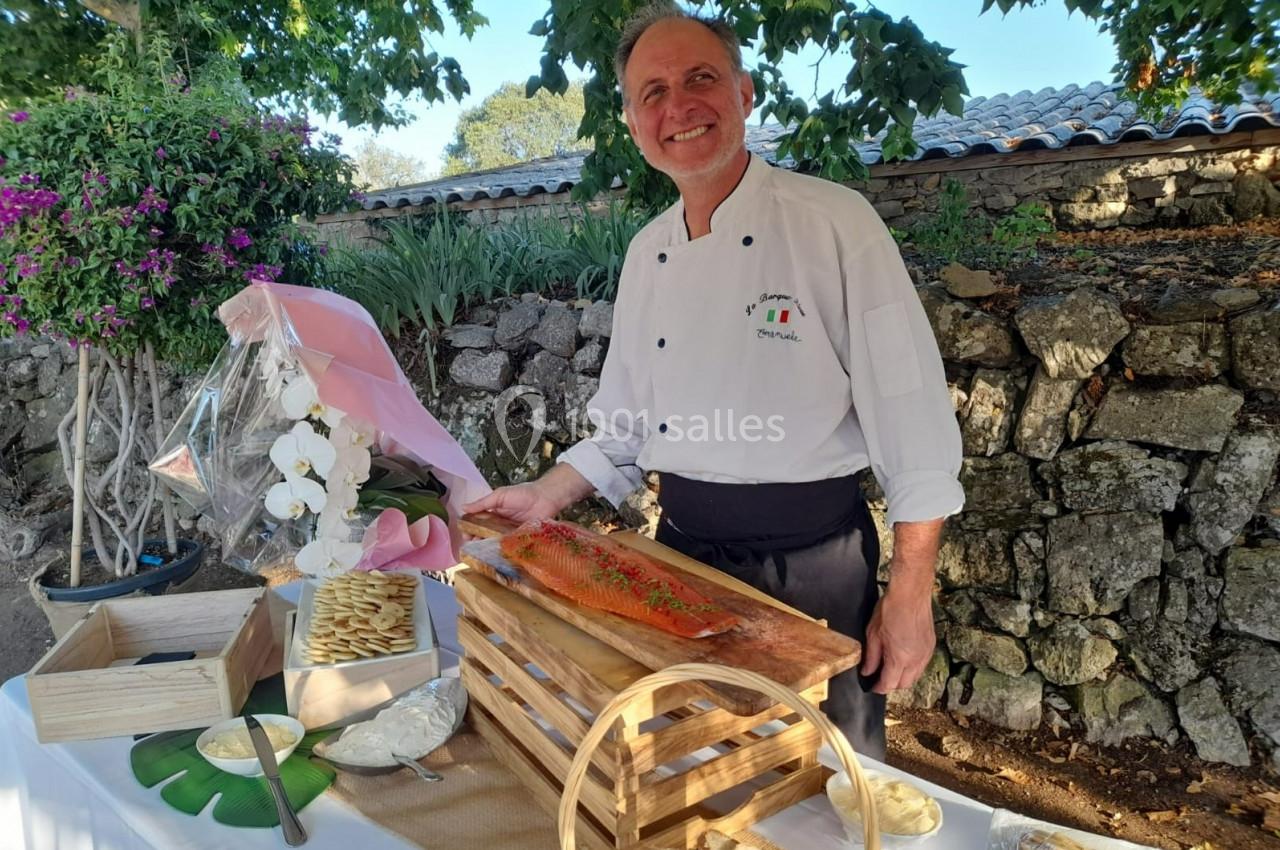 Un chef souriant présente un saumon fumé sur une table dressée en extérieur, entourée de fleurs et de pierres.