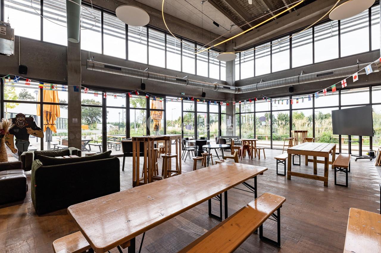 Salle lumineuse avec de grandes baies vitrées, tables en bois, chaises et décorations suspendues.