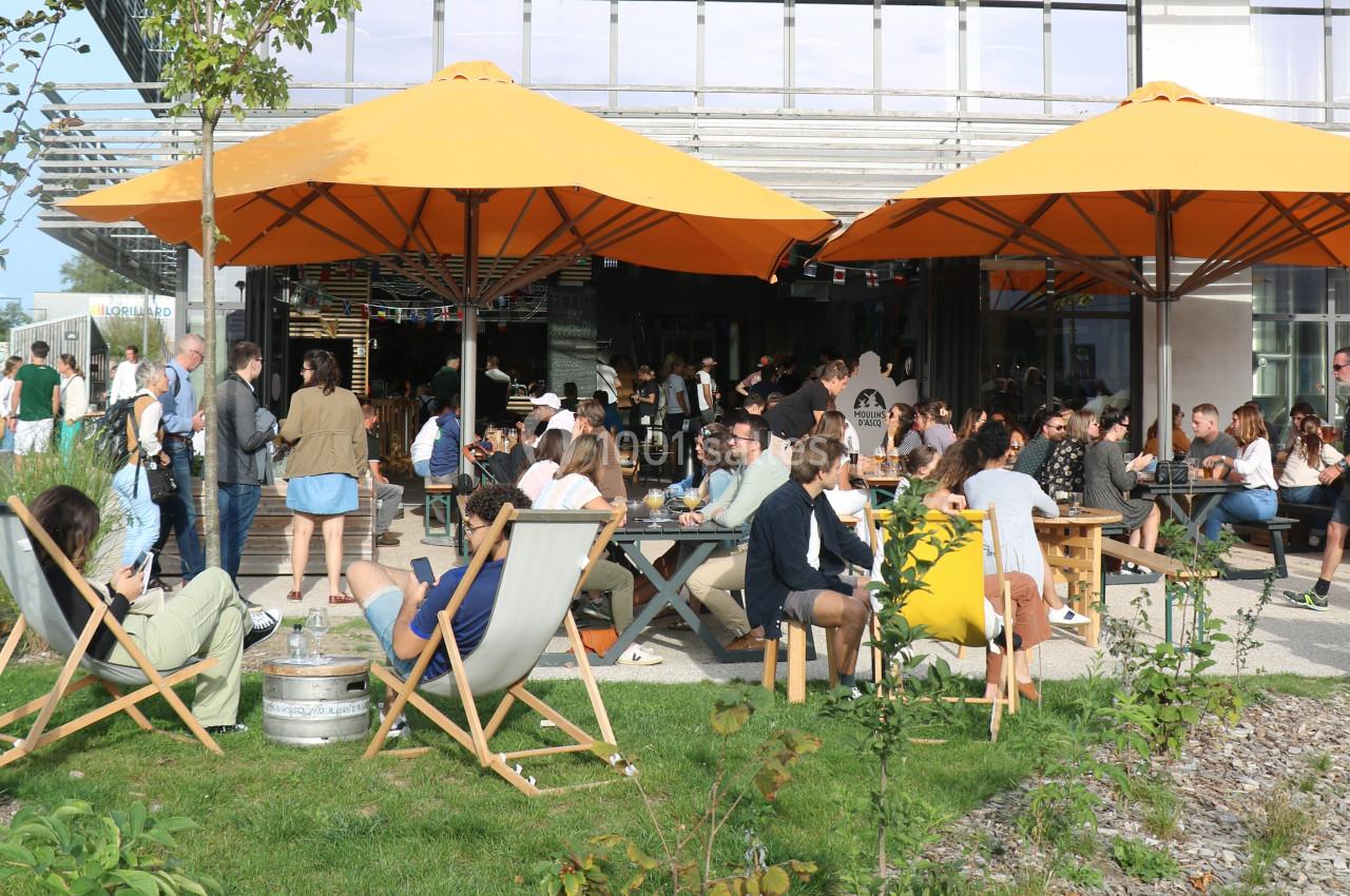 Terrasse animée avec des tables et chaises longues, des personnes profitant du soleil sous des parasols orange.