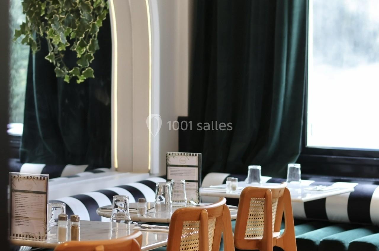Salle de restaurant lumineuse avec des chaises en bois, banquettes vertes, tables dressées et plantes suspendues.