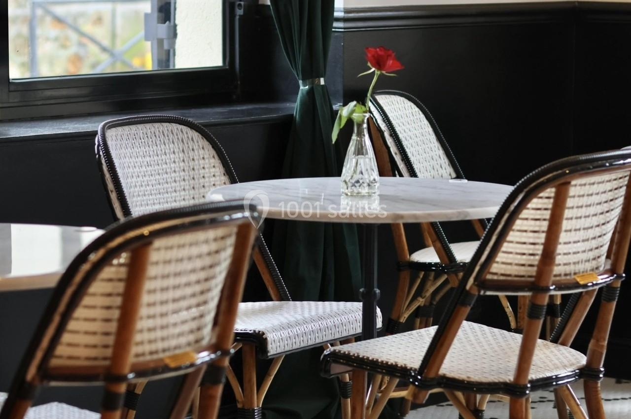 Table en marbre entourée de chaises en rotin dans un café au sol carrelé, près d'une fenêtre avec rideaux verts.