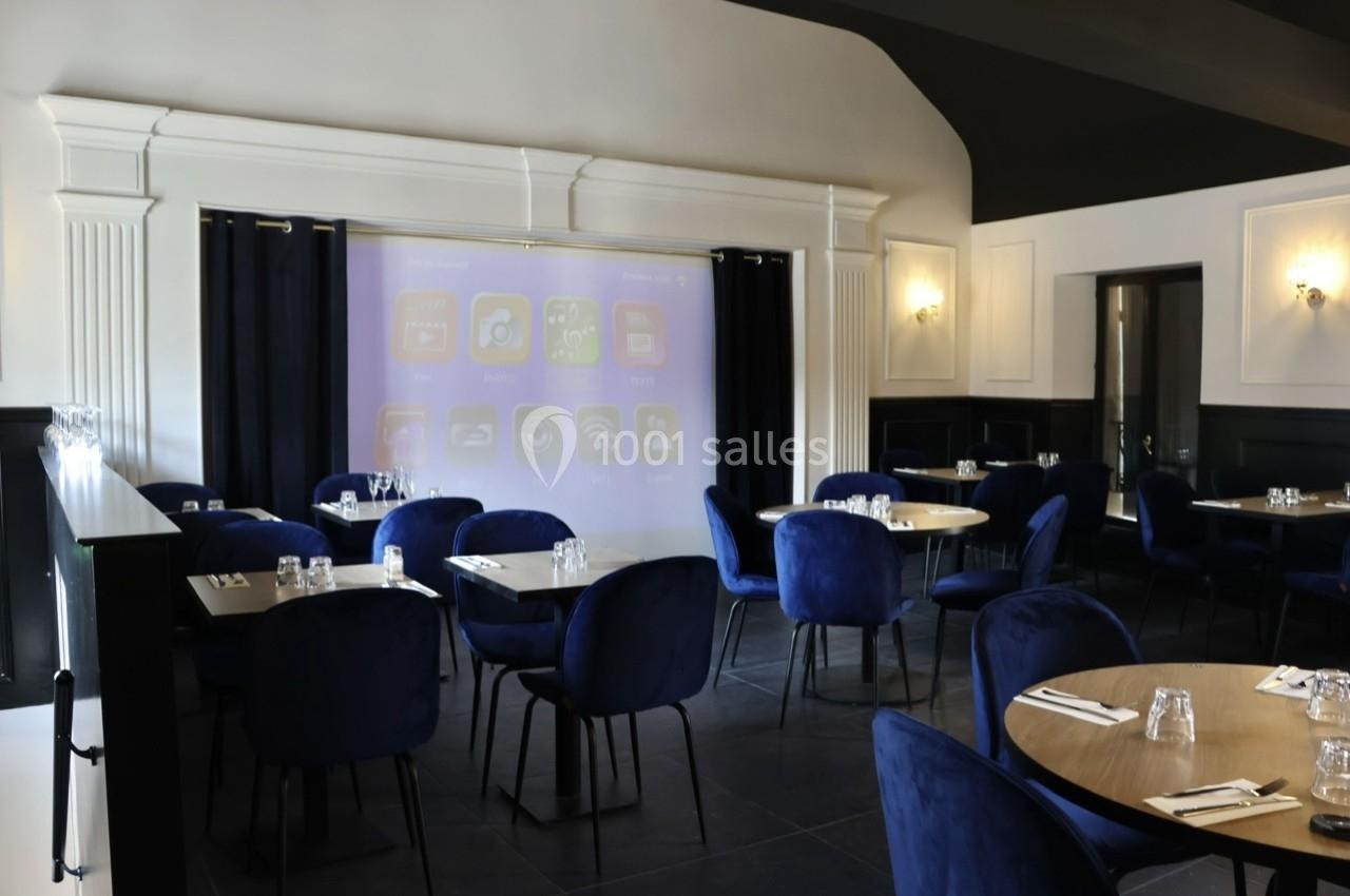 Salle de restaurant moderne avec tables rondes, chaises bleues et un écran de projection en arrière-plan.