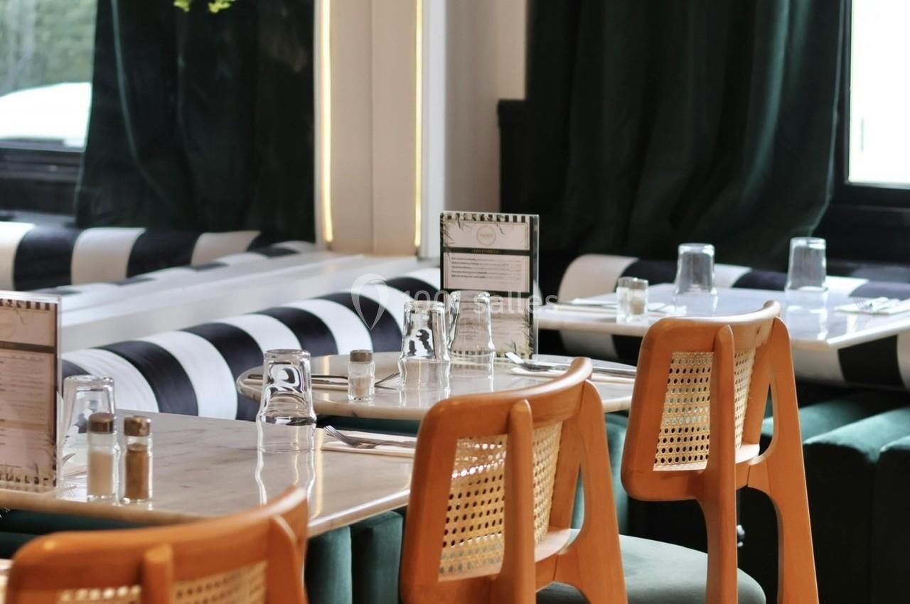 Salle de restaurant élégante avec tables en bois, chaises en cannage et banquettes vertes devant des rideaux sombres.