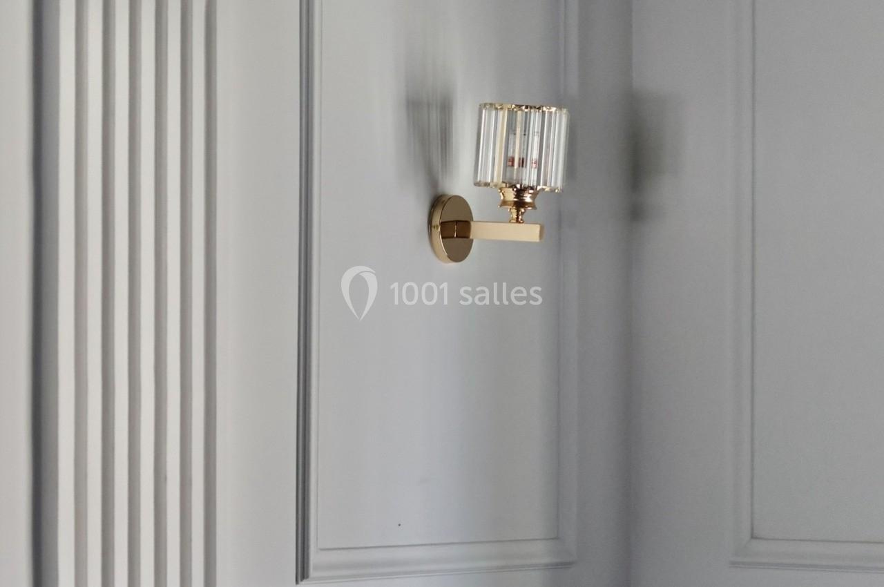 Applique murale dorée avec abat-jour en verre, fixée sur un mur blanc orné de moulures et contrasté par un soubassement noir.